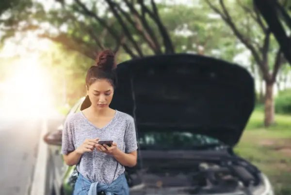 A woman stands beside her car on a tree-lined road, looking at her phone with the hood propped open behind her.