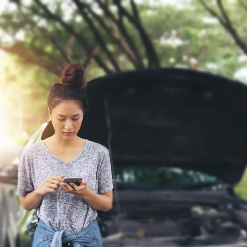 A woman stands beside her car on a tree-lined road, looking at her phone with the hood propped open behind her.