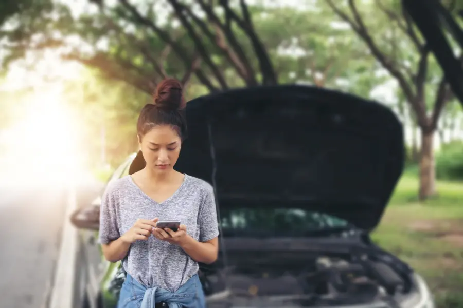 A woman stands beside her car on a tree-lined road, looking at her phone with the hood propped open behind her.