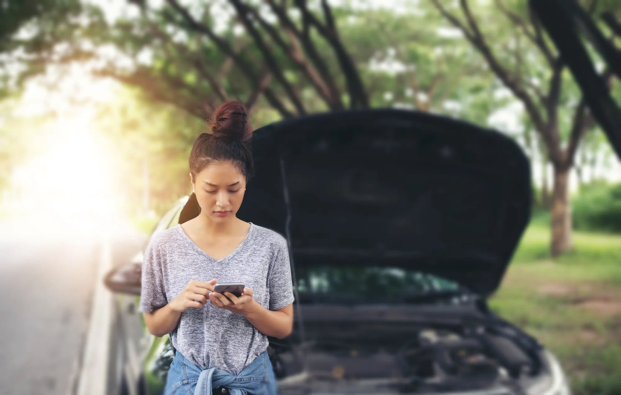 A woman stands beside her car on a tree-lined road, looking at her phone with the hood propped open behind her.