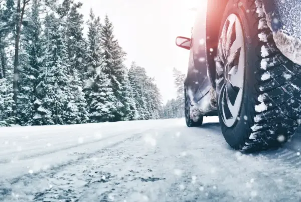 Close-up of a car's snow-covered tire on a snowy road lined with snow-laden evergreen trees.